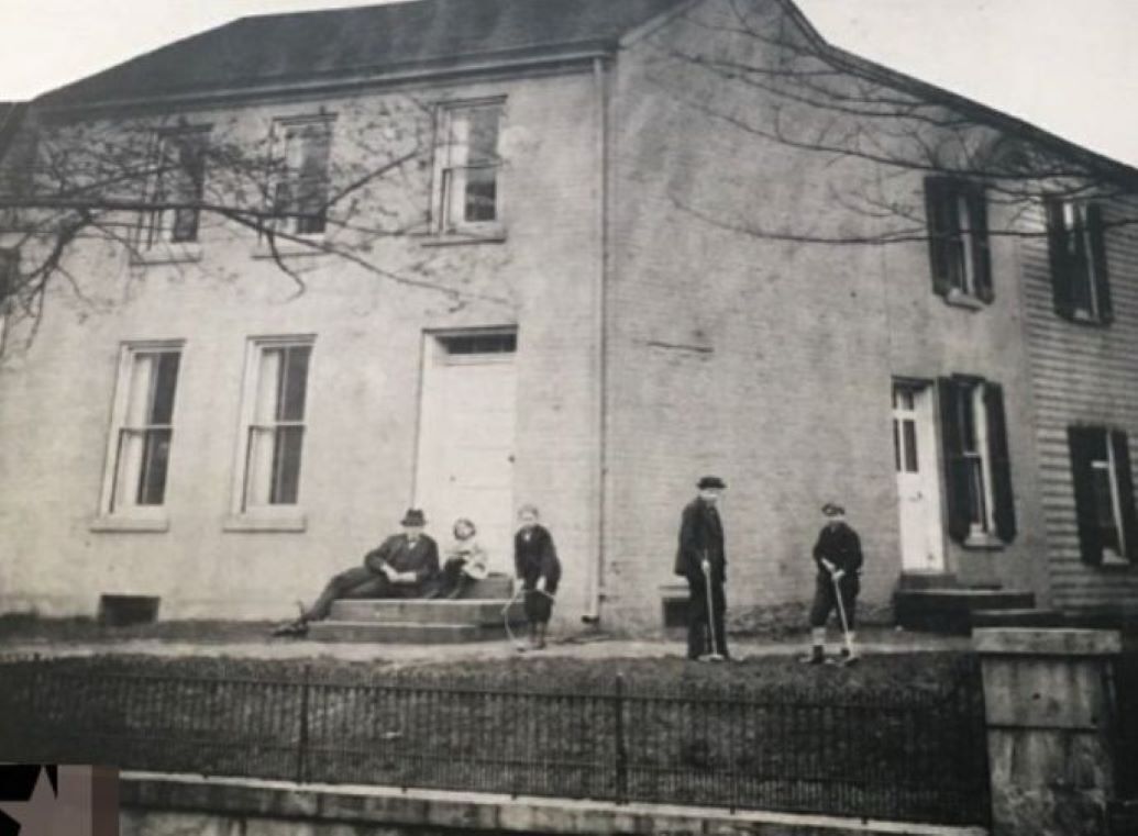 a vintage image of an old brick house with people playing croquet in the yard, a boy hoop rolling, and a man and child sitting on the front door stoop